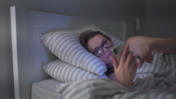 Mujer con gafas utiliza un teléfono inteligente mientras está acostado en la cama. Ella se queda dormida en el proceso porque está muy cansada
 - Metraje, vídeo