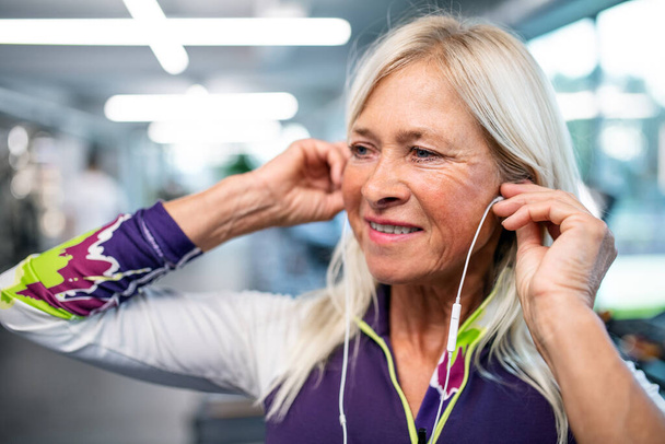 Una mujer mayor con auriculares en el gimnasio descansando después de hacer ejercicio
. - Foto, imagen