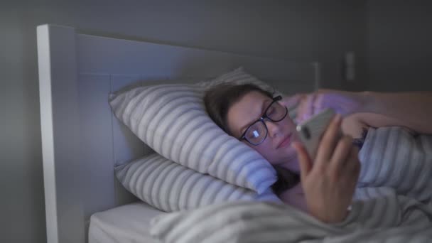 Mujer con gafas utiliza un teléfono inteligente mientras está acostado en la cama. Se frota los ojos, porque está cansada y somnolienta. Concepto de adicto móvil o insomnio
. - Metraje, vídeo