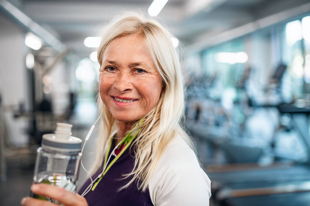 A senior woman with earphones and water bottle standing in gym. - Valokuva, kuva