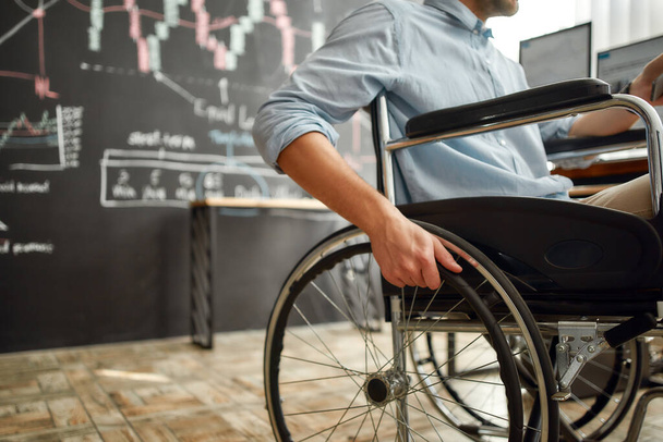 Cropped photo of male office worker in a wheelchair working in the modern office. Disabled man holding a hand on wheel of a wheelchair while sitting at his workplace - Photo, image