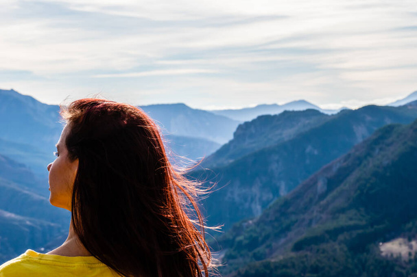 A close-up back view of a young redhead woman with her long hair fluttering in front of the mountain valley landscape from behind on a sunny windy day - Photo, Image