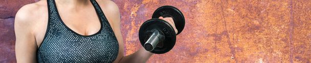 A young slim athletic girl in sportswear performs a set of exercises on a grunge rusty backgrounds. Performs exercises with dumbbells. Fitness and healthy lifestyle. - Foto, imagen