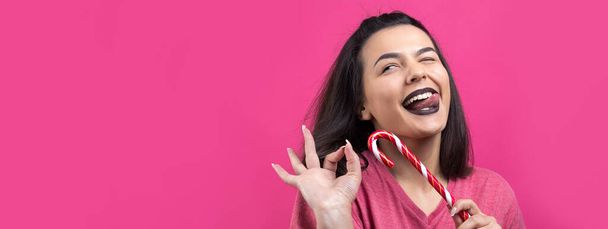 Πορτρέτο του όμορφη γλυκιά χαρούμενη γυναίκα με ίσια καστανά μαλλιά προσπαθεί να δαγκώσει κόκκινο ζαχαροκάλαμο καραμέλα Χριστούγεννα. - Φωτογραφία, εικόνα