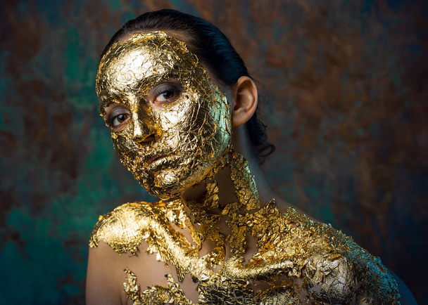 Girl with a mask on her face made of gold leaf. Gloomy studio portrait of a brunette on an abstract background. - Fotoğraf, Görsel