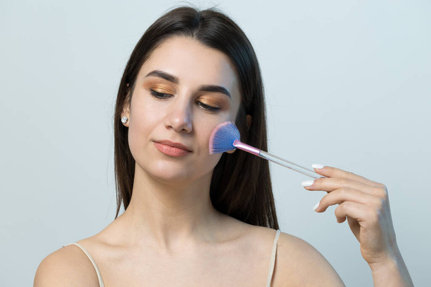 Close-up of a young girl in a light top on a white background making a facial make-up. A pretty woman holds a cosmetic brush near her face and smiles. - Φωτογραφία, εικόνα
