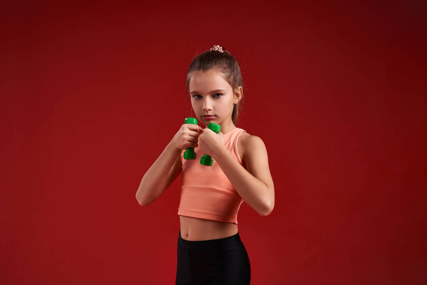 Be prepared. A cute kid, girl is engaged in sport, she is looking at camera while exercising with dumbbells. Isolated on red background. Fitness, training, active lifestyle concept. Horizontal shot - Fotó, kép