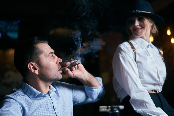 The man-gangster and the beautiful senior woman smoking cigar in a bar - Photo, Image