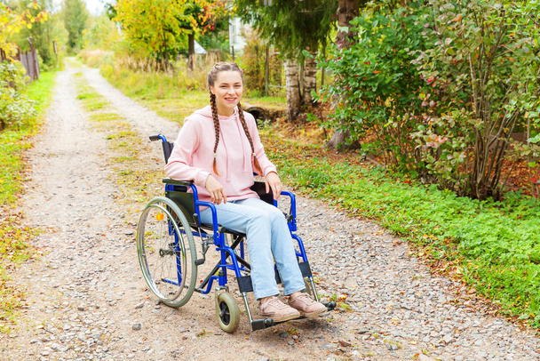 Jovem mulher deficiente feliz em cadeira de rodas na estrada no parque hospitalar à espera de serviços de pacientes. Menina paralisada em cadeira inválida para pessoas com deficiência ao ar livre na natureza. Conceito de reabilitação. - Foto, Imagem