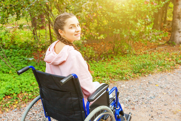 患者サービスを待っている病院の公園の道路上の車椅子の若い幸せなハンディキャップの女性。自然の中で屋外の障害者のための無効な椅子に麻痺した女の子.リハビリテーションの概念. - 写真・画像