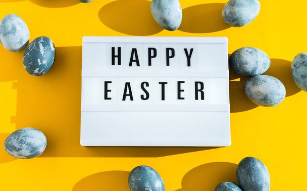 Blue textured painted Easter eggs and a luminous decorative board with the letters Happy Easter on a bright yellow holiday background backdrop. Hard light. Long shadow. - Foto, afbeelding