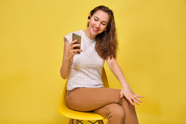 A girl in a T-shirt on a yellow background with wireless headphones takes a selfie and shares a photo on social networks. - Foto, Imagem