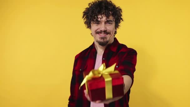 A man gives a red gift for Valentines Day lovers or International Womens Day. Birthday surprise. Handsome happy european man with beard in shirt smiling isolated on yellow background. Slow motion. - Video, Çekim