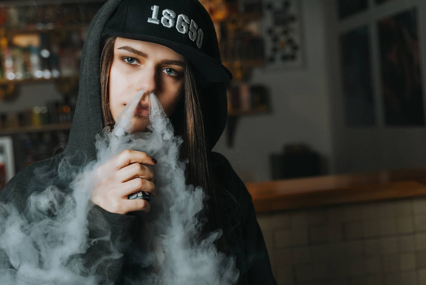 Una joven guapa con gorra fuma un cigarrillo electrónico en la tienda de vapores. Estilo hip-hop. Primer plano.
. - Foto, imagen
