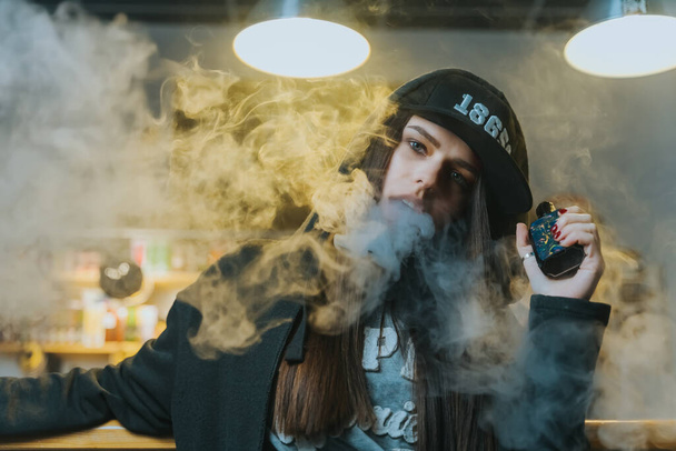 Young pretty woman in cap smoke an electronic cigarette at the vape shop. Hip-hop style. Closeup. - Фото, изображение