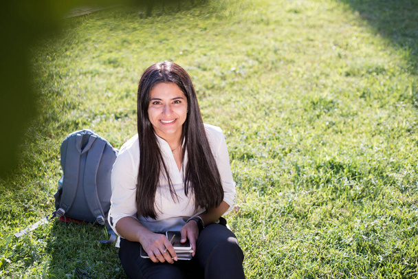 Індійська дівчинка, студентка, усміхається, сидить на зеленій траві з рюкзаком і блокнотом, пише: - Фото, зображення