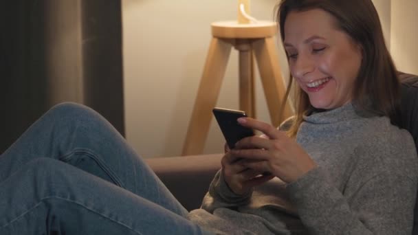 Smiling woman lying on the couch in a cozy room and using smartphone for surfing internet in the evening. Relaxation and lifestyle technology. - Кадри, відео