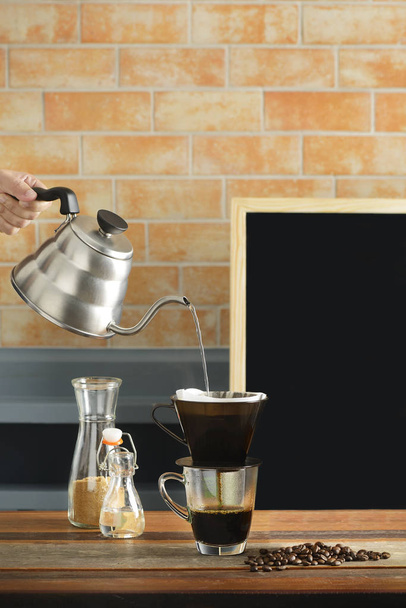 Рука тримає чайник для крапельного заварювання кави, наливаючи гарячу воду на кавові зерна та фільтруючи на чашку кави в кав'ярні, цегляній стіні та дошці на задньому плані
. - Фото, зображення