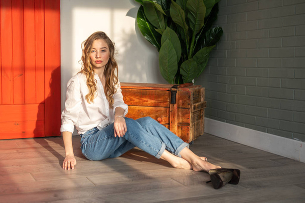 Young beautiful woman sitting on the floor with bare feet next to wooden box and green plant - Photo, Image