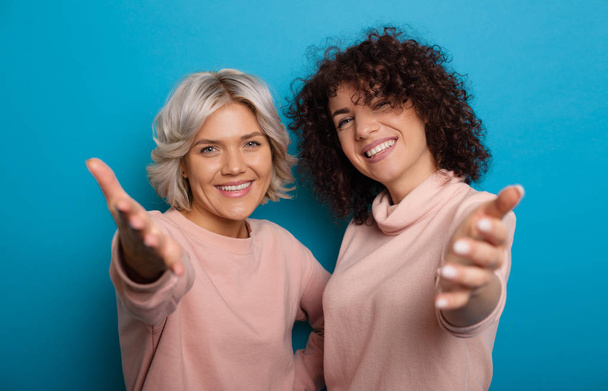 Two smiling caucasian ladies with curly hair pointing to you as an invitation on blue background - Fotografie, Obrázek