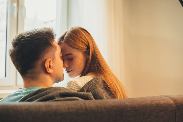 Veselá dáma s rudými vlasy a pihami v držení svého přítele se těší jeho polibky, zatímco je pokryta přikrývkou - Fotografie, Obrázek