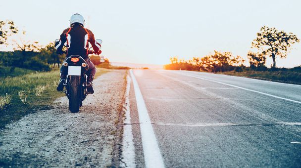 Achteraanzicht van een blanke man goed uitgerust beginnen te rijden een motorfiets met een zonsondergang op de achtergrond - Foto, afbeelding