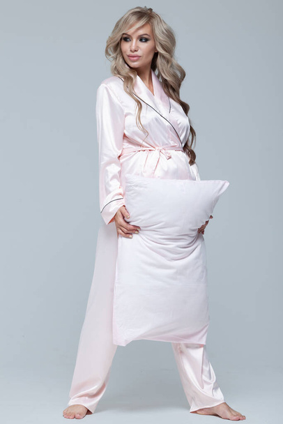 Young beautiful pregnant woman in pink pajamas with pink pillow. The concept of stylish pregnancy and beautiful sleep. On a gray uniform background. Copy paste. Full length. - Foto, Bild