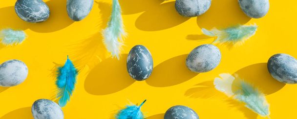 Blue textured colored Easter eggs and chicken bird feathers on a bright yellow holiday background backdrop. Spring holiday concept. Copyspace. Flat lay. Top view. Hard light. Long shadow. - Photo, Image