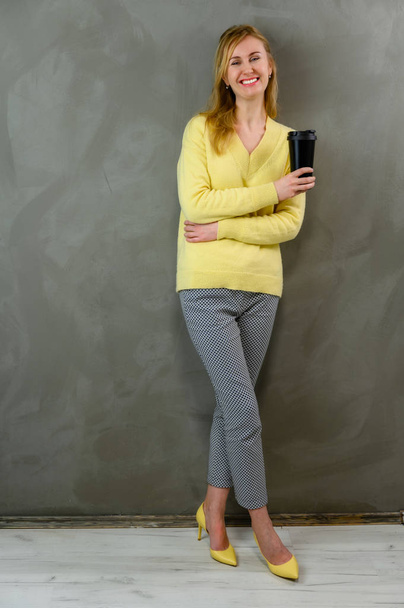 Joyful blond Caucasian woman in a yellow sweater and gray trousers stands right with coffee in her hands on a gray background near the wall in full length - Foto, Imagen