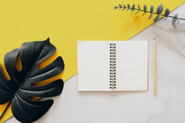 Tropisches grünes Blatt und Notizbuch mit Bleistift auf gelber Wand und weißem Marmor Textur abstrakten Hintergrund. - Foto, Bild
