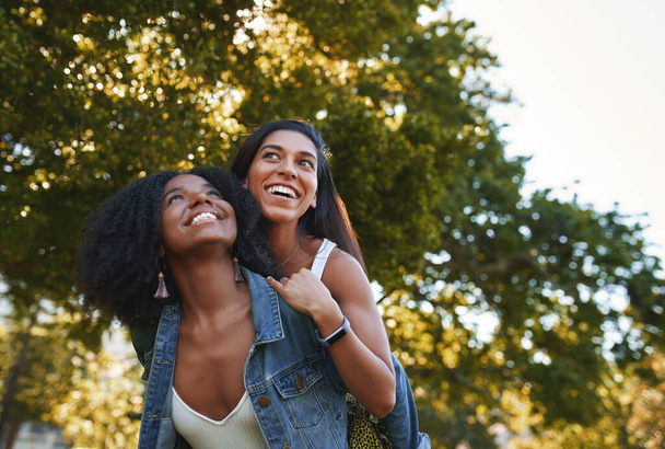 Portrait of a smiling young african american woman giving a piggyback ride to her female best friend in the park on a sunny day - group of college students laughing and having fun on campus - Фото, зображення
