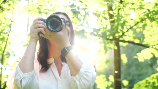 Close-up view of a young girl who takes a photo with a professional camera on the background of a bright blinding sun. A shot near the face of a beautiful smiling girl who takes pictures of nature. - Materiał filmowy, wideo
