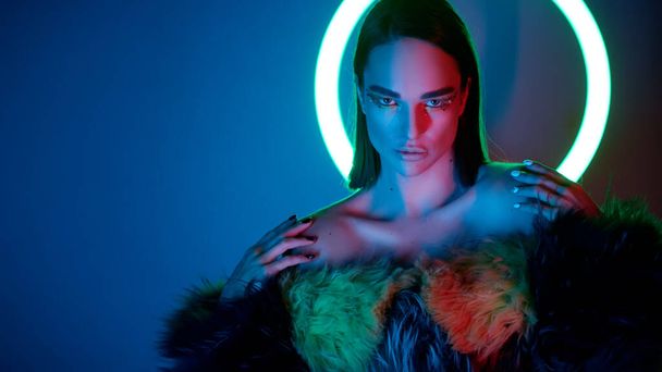 beautiful model with bright makeup is boldly looking at the camera against the background of light lamps in a fur coat. High end retouch. - Foto, Imagem