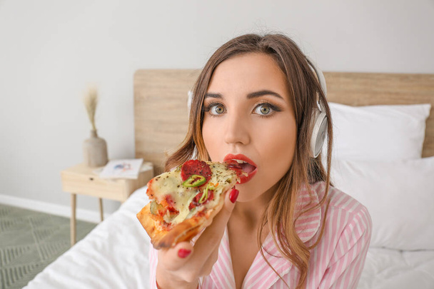 Утром красивая молодая женщина ест вкусную пиццу и слушает музыку в спальне
 - Фото, изображение