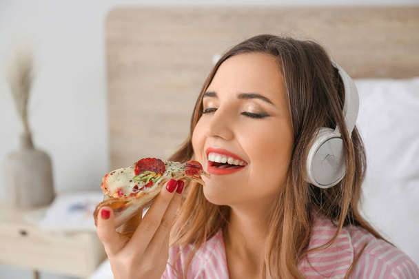 Morning of beautiful young woman eating tasty pizza and listening to music in bedroom - Фото, изображение