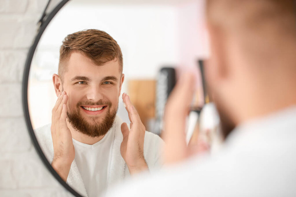 Handsome young man applying cream on his face near mirror in bathroom - Фото, зображення