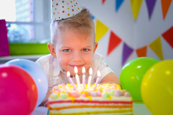 Blonde caucasian little boy looking at candles on birthday rainbow cake, making a wish before blows them out at birthday party. Colorful background with balloons - Foto, Imagen