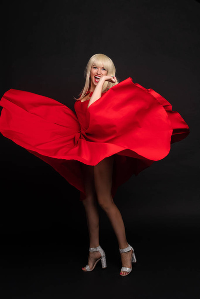 Мода усміхнена жіноча модель позує граючи з махаючим червоним сексуальним платтям, дивлячись на повний знімок камери
 - Фото, зображення