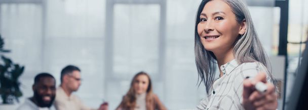 Panoramaaufnahme einer lächelnden asiatischen Geschäftsfrau, die mit Filzstift auf ihre multikulturellen Kollegen im Hintergrund zeigt  - Foto, Bild