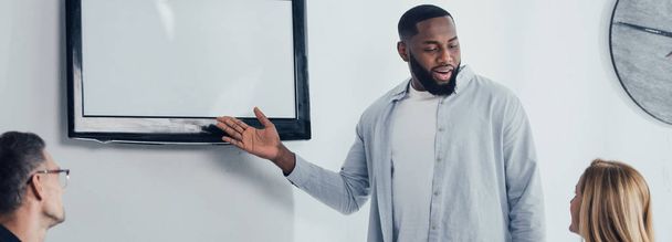 панорамний постріл усміхненого афроамериканського бізнесмена, який вказує рукою на ТВ і розмовляє з колегами.  - Фото, зображення
