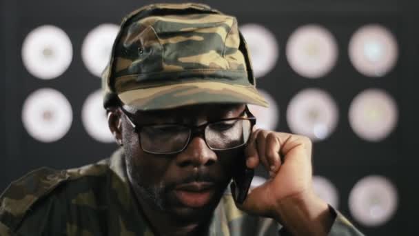Bearded mature black guy dressed in military clothes and cap, black glasses asits at desk and speaks on phone in dark modern office room with blurred background. - Séquence, vidéo