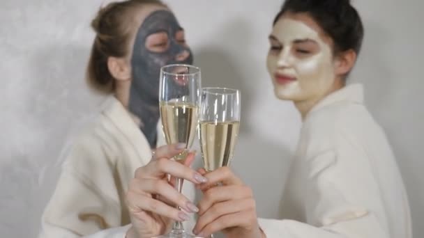 Cheerful beautiful caucasian young female best friends wearing white bathrobes holding glasses with champagne laughing at camera, two happy playful ladies gather on spa pajama party or bridal shower - Footage, Video