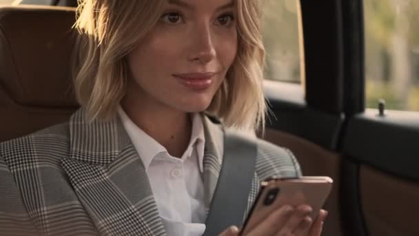 Close up view of Happy blonde business woman in coat using smartphone and looking away while sitting in car - Footage, Video