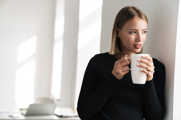 Blonde business woman in office indoors drinking hot tea. - Foto, afbeelding