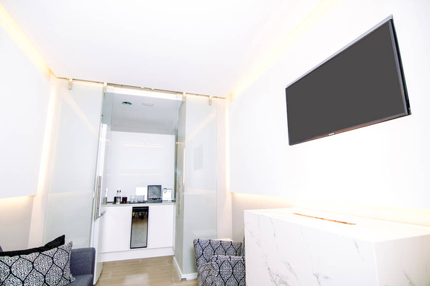 Современный зал ожидания с белыми панелями и телевизором в стене и технологическим мраморным камином. В конце зала есть небольшая кухня. - Фото, изображение