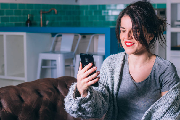 Smiling young woman looking at her smartphone screen while sitting on a couch at home - Foto, imagen