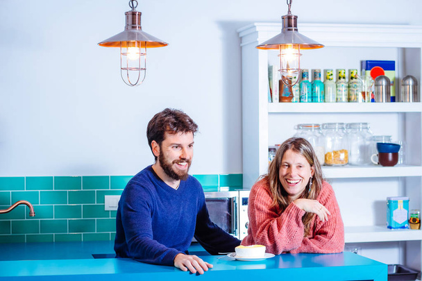 Ευτυχισμένο νεαρό ζευγάρι γελάει καθισμένο σε μια μοντέρνα κουζίνα ενώ μοιράζεται ένα κέικ λεμονιού. Η έννοια του Αγίου Βαλεντίνου. - Φωτογραφία, εικόνα