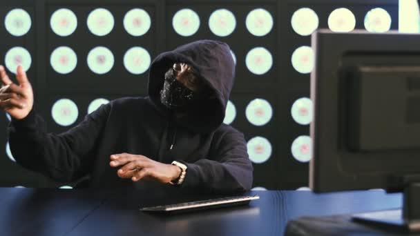 Africano-americano maturo maschio computer di sicurezza hacker indossando maschera viso nero e felpa è seduto in camera moderna scura al desktop, mettendo via il suo smartphone e inizia a scrivere attivamente sulla tastiera. - Filmati, video