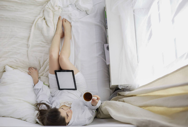 朝、ベッドに座りながらタブレットやコーヒーを飲んでいる女性の肖像画 - 写真・画像