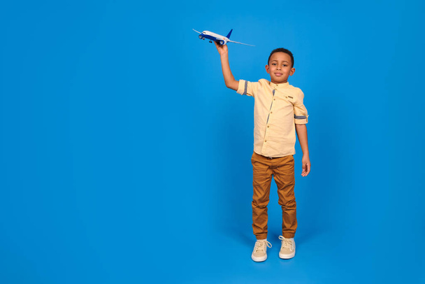 Happy boy African American in brown pants and a light shirt plays with a toy plane, imagines him flying in a plane against a blue background. Family travel and tourism, summer holidays consept. - Photo, image